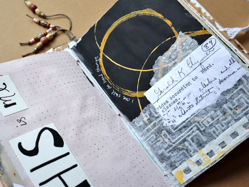 Pages in a handmade art journal showing a collage that includes a piece of an old test, a textured stencil of an electric circuit, and stamped circles in a mustard color on a black background.