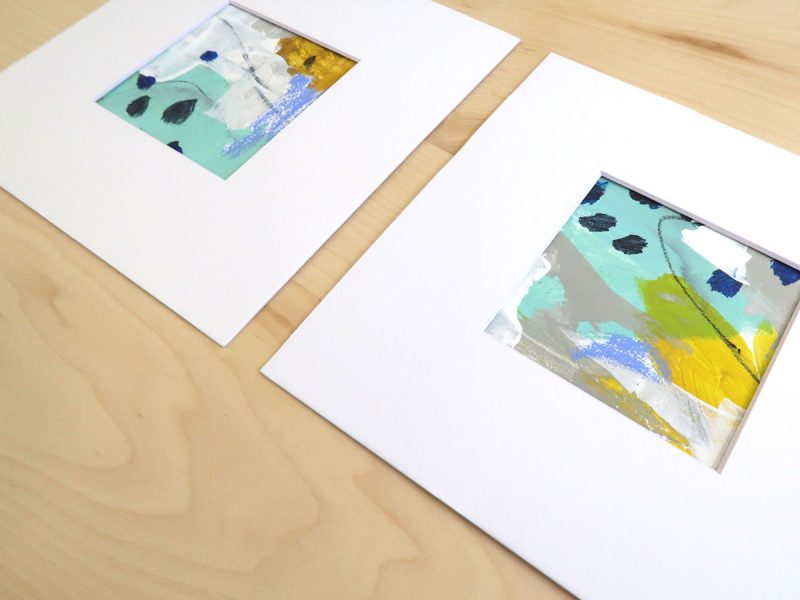Two small, square abstract paintings in turquoise, yellow, gray and white shown matted on a wood background