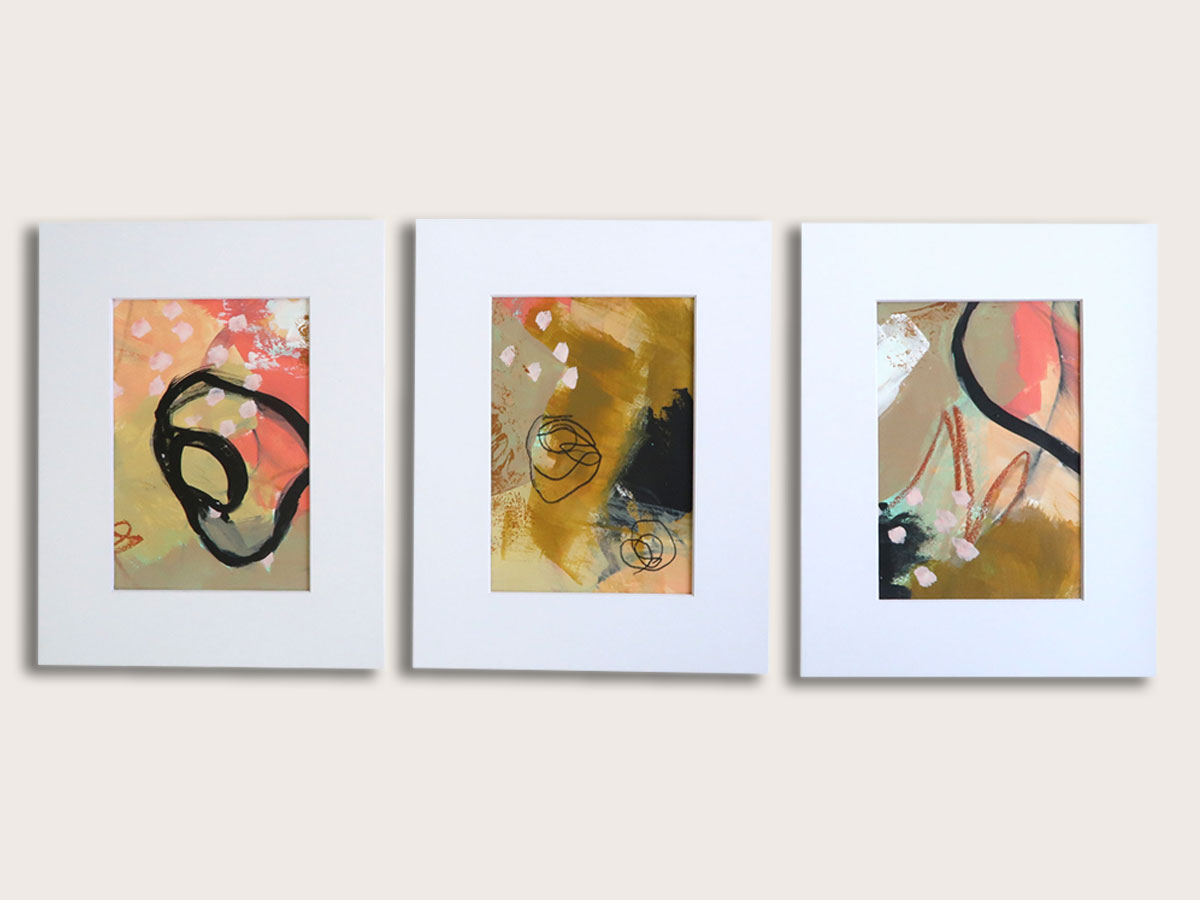 Set of three colorful abstract paintings with backgrounds in tones of pink, gold, beige, and black. Thick black lines form irregular shapes and all three have a section of light pink dots. Each has a bright white mat.