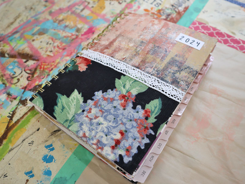 2024 spiral bound planner with custom cover design with painted paper on top, floral fabric on the bottom, and a crochet ribbon